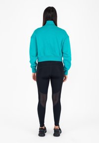 Felpa corta turchese con colletto alto, abbinata a leggings neri con pannelli in rete e scarpe da ginnastica nere.