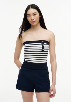 Giovane donna con lunghi capelli scuri che indossa un top senza spalline a righe blu navy e bianco e pantaloncini a vita alta blu navy con dettagli a bottoni.