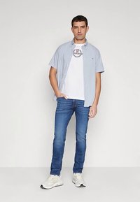 Lyseblå kortærmet skjorte med knapper over en hvid t-shirt med logo, kombineret med blå jeans og hvide sneakers. Neutral baggrund.