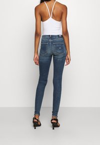 Úzké džíny v tmavě modrém denimovém materiálu, s vysokým pasem, dvěma zadními kapsami a hladkou texturou, kombinované s bílým topem na špagetových ramínkách.