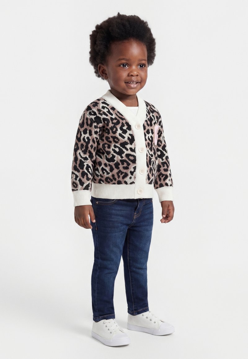 Jeune enfant aux cheveux bouclés portant un cardigan à imprimé léopard, un jean bleu foncé et des baskets blanches, debout et souriant.