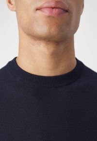 Gros plan sur le bas du visage et le cou d'une personne portant un pull à col rond bleu foncé sur un fond clair uni.