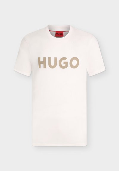 Бяла тениска с къси ръкави и кръгло деколте с бежов надпис "HUGO" на гърдите и червен етикет отвътре на деколтето.