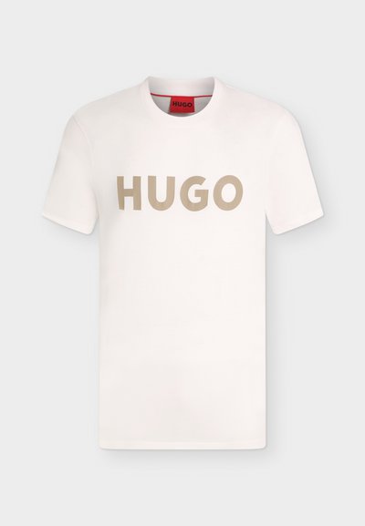Бяла тениска с къси ръкави и кръгло деколте с бежов надпис "HUGO" на гърдите и червен етикет отвътре на деколтето.