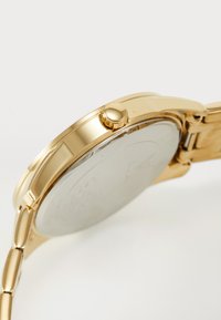 Guldtonet metal armbåndsur med en rund sag, en trykkrone og et sammenkoblet armbånd. Bagsiden har en glat metallisk overflade.