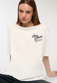 Biała, oversize'owa koszulka z krótkim rękawem i okrągłym dekoltem, z czarnym nadrukiem zawierającym napis "Ciao Amore" oraz wzór serca.
