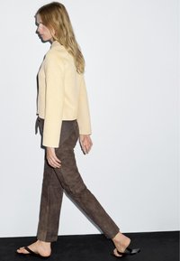 Beige korte jas met lange mouwen, gecombineerd met bruine suède broek. Model loopt in zwarte slippers tegen een effen witte achtergrond.