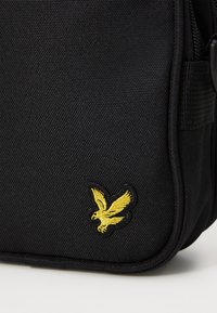 Zwarte stoffen tas met een gestructureerde oppervlakte, voorzien van een gele geborduurde adelaarlogo aan de voorkant. Inclusief zwarte ritsen en piping.