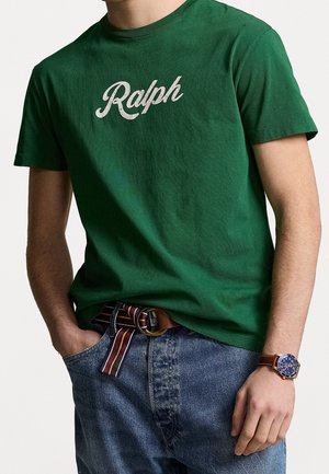 Homme portant un t-shirt vert "Ralph", un jean bleu avec une ceinture en tissu rayé, et une montre-bracelet bleue avec un bracelet en cuir marron.
