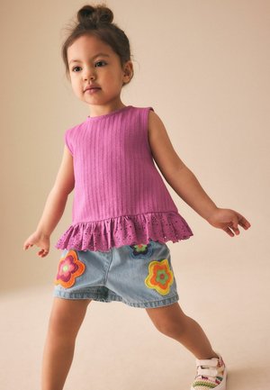 Kleinkind mit einem Dutt, das ein ärmelloses rosa Oberteil mit Rüschen und Jeansshorts trägt, die mit bunten Blumenapplikationen verziert sind.