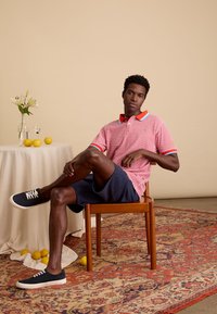 Rood-wit geruite polo met blauwe en rode accenten, marineblauwe shorts, donkere sneakers, zittend op een houten stoel tegen een neutrale achtergrond.