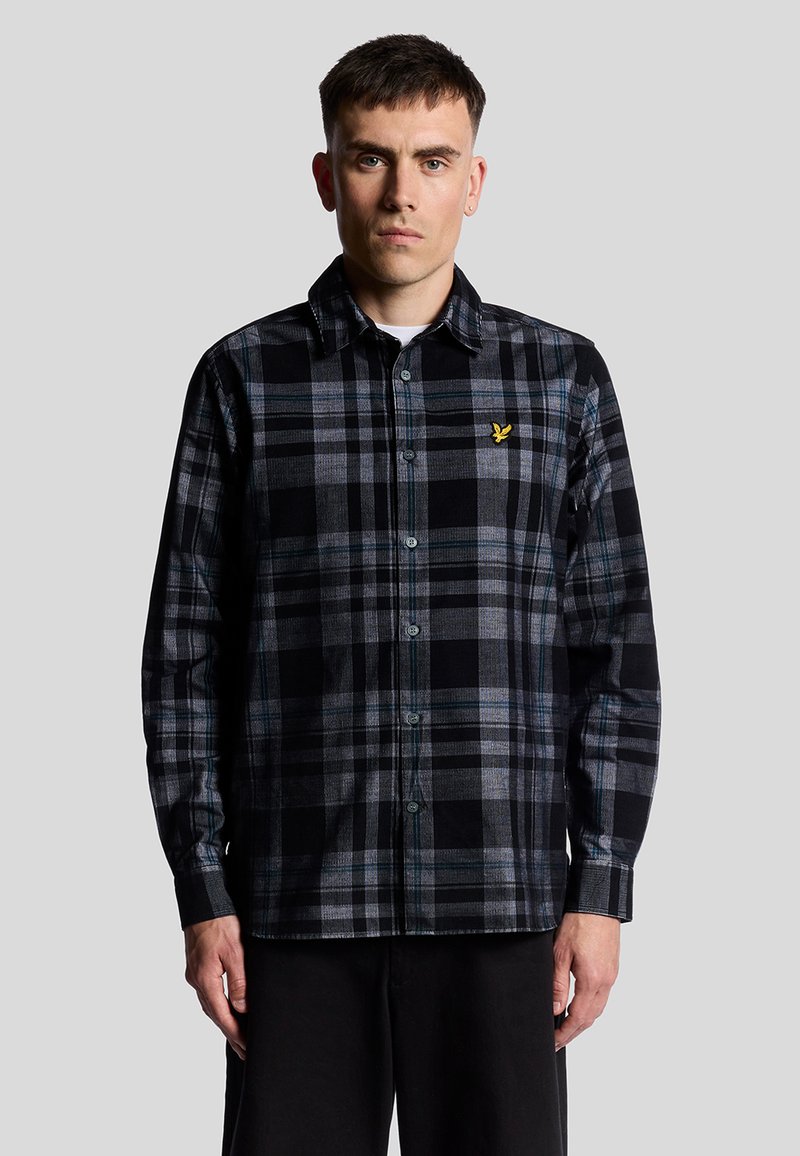 Camisa de franela negra y gris con patrón de cuadros, diseño de botones, acentos en azul y blanco en contraste, y un pequeño logo amarillo en el pecho.