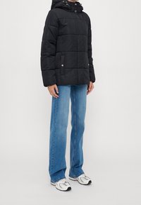 Svart quiltad puffjacka med hood, som har två framfickor, detaljer i guld och en avslappnad passform, bärs över blå jeans.