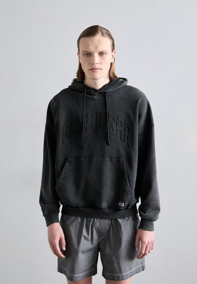 Hanorac negru supradimensionat cu text embossat, având un buzunar frontal și glugă cu șnur, purtat cu pantaloni scurți gri pentru un look casual.