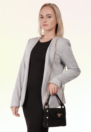 Femme aux cheveux blonds portant un blazer gris et une robe noire, tenant un sac à main noir avec un emblème d’abeille doré et une bandoulière chaîne.