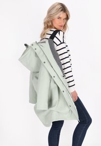 Giacca impermeabile verde menta chiaro con rivestimento grigio, chiusura a zip, bottoni a pressione e cappuccio, indossata sopra una maglietta a maniche lunghe a righe.