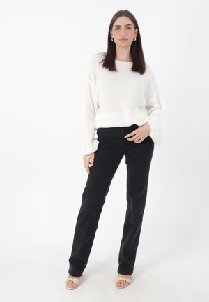 Witte gebreide trui met brede halslijn, gecombineerd met zwarte broek. De outfit wordt aangevuld met open teen beige sandalen. Effen witte achtergrond.