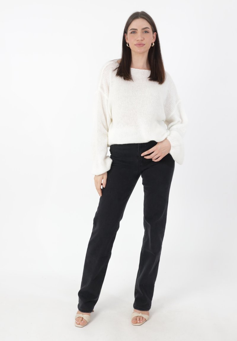 Witte gebreide trui met brede halslijn, gecombineerd met zwarte broek. De outfit wordt aangevuld met open teen beige sandalen. Effen witte achtergrond.