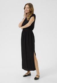 Black maxi dress with short sleeves, elastic waist, side slits, and a smooth texture. Worn with flat black shoes.