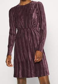 Vestido de terciopelo púrpura de manga larga con pliegues verticales, llevado por una persona de piel oscura que lleva un anillo dorado en la mano derecha.