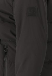 Svart isolerad jackärm med en dragkedjficka och en texturerad etikett med ordet "WHISTLER" och geometrisk design.
