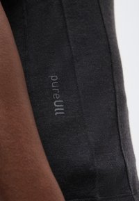 Prim-plan al mânecii din țesătură închisă la culoare, cu textul „pureWool” imprimat în gri deschis pe brațul cu piele închisă al unei persoane.