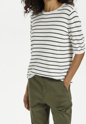 Personne portant une chemise blanche à rayures horizontales noires et un pantalon vert olive avec un détail de poche cloutée, main dans la poche.