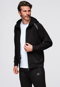 Sudadera con cremallera negra y capucha, con un tejido suave y detalle en texto blanco en el hombro. Llevada sobre una camisa clara.
