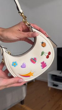 Weißes Lederhandtasche mit einer geschwungenen Form, die mit verschiedenen bunten Aufklebern verziert ist, darunter ein Marienkäfer, Kirschen, Blumen und ein Sternakzent.