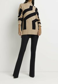 Brede trui in beige met zwarte geometrische strepen, hoge halslijn, geribde manchetten en zoom. Gecombineerd met zwarte flared broeken en puntige hakken.