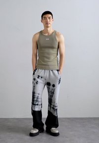 Pantaloni de trening gri tie-dye cu accente negre, asortați cu un top de culoare verde măsliniu cu un logo. Modelul stă într-un fundal simplu.