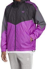Chaqueta cortavientos morada y gris con capucha, parte delantera con cremallera y puños elásticos. Presenta paneles en contraste y bolsillos laterales.