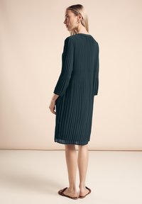 Dunkelgrünes, plissiertes Kleid mit drei Viertelärmeln und rundem Ausschnitt, das einen Knopfverschluss am Rücken hat. Glatte Textur mit fließendem Design.