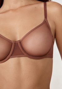 Sujetador marrón de malla con aros, copas lisas y semi-transparentes, tirantes ajustables y una costura central para estructura y soporte.