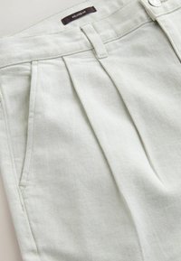 Detailansicht einer hellgrauen Hose mit entspanntem Schnitt, die Vordertasche, Gürtelschlaufe, Falte und ein "Relaxed Fit"-Etikett nahe dem Bundknopf zeigt.