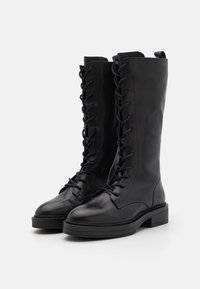 Botas de cuero negras de altura hasta la rodilla, con cordones, textura lisa, punta redondeada y suela de goma plana. Cadenas de ojales suben por el frente.