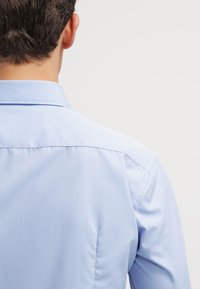 Cămașă de culoare albastru deschis, cu guler clasic și textură netedă, țesută. Vederea din spate arată detalii de cusătură și un design ajustat.