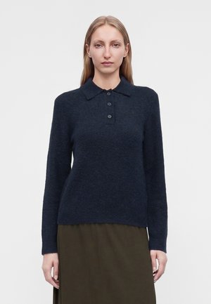 Femme aux longs cheveux blonds portant un pull bleu marine foncé à manches longues avec des boutons et une jupe vert olive sur un fond uni.