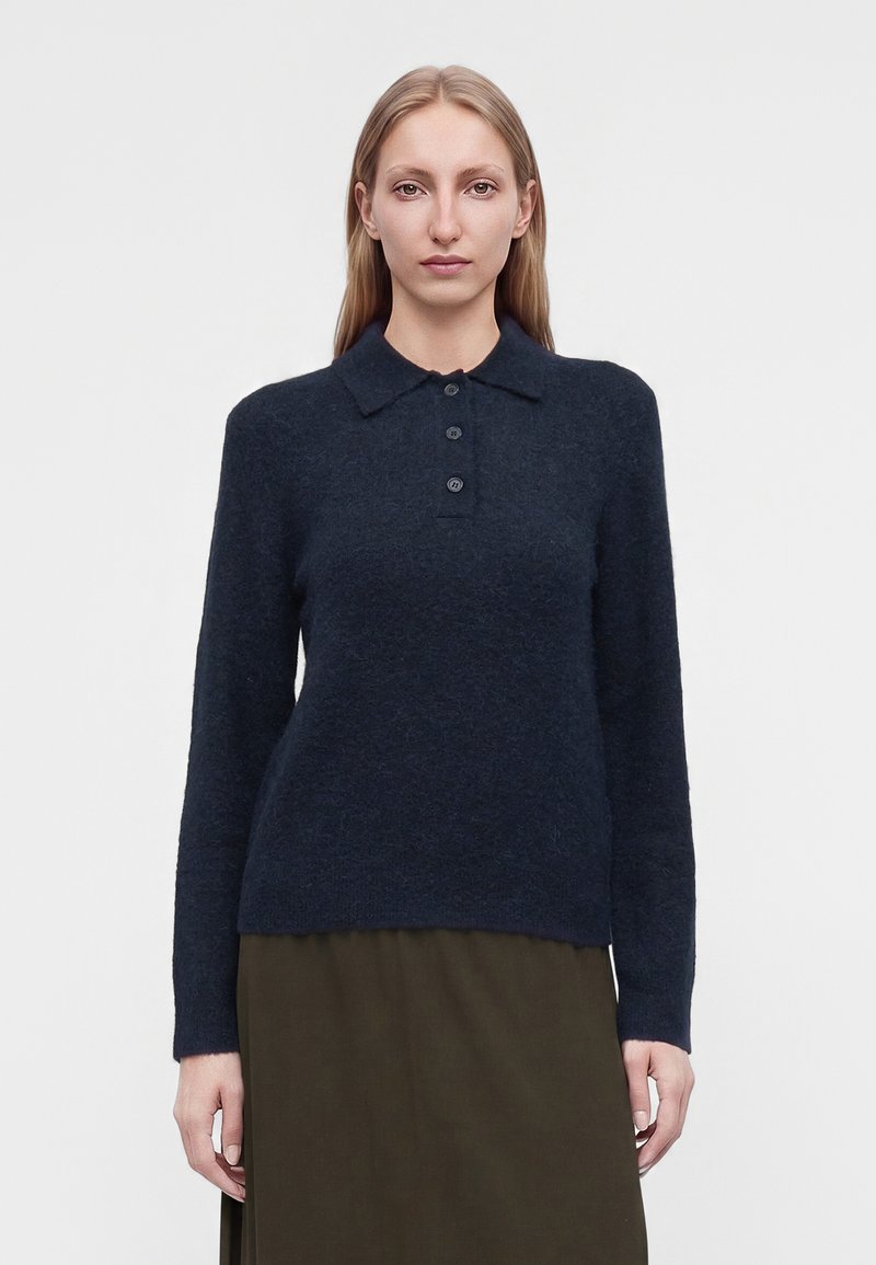 Femme aux longs cheveux blonds portant un pull bleu marine foncé à manches longues avec des boutons et une jupe vert olive sur un fond uni.