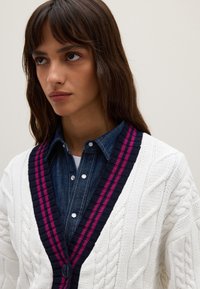 Weißer Strickcardigan mit tiefblauem und magentafarbenem gestreiften Saum, kombiniert mit einem Jeanshemd mit Knopfleiste.