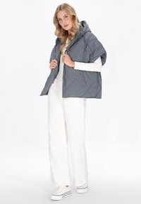 Vest puffer griș cu o țesătură matlasată, cu glugă, mâneci scurte, fermoar frontal și detalii cu prindere, purtat peste un tricou alb cu mânecă lungă și pantaloni albi cu picioare late.
