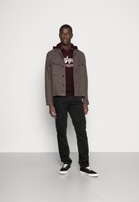 Grå knappejakke over en maroon hettegenser, svarte cargobukser og svarte sneakers med hvite såler. Glatt tekstur, avslappet design.