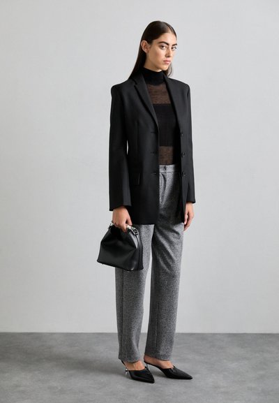 Schwarzer taillierter Blazer über einem durchsichtigen schwarzen Top, graue Hahnentritt-Hosen, schwarze spitze Schuhe und eine strukturierte schwarze Handtasche.