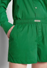 Personne vêtue d'une tenue verte à manches longues avec un short assorti à taille élastique, main dans la poche gauche, étiquette indiquant "DESIGNERS, REMIX."