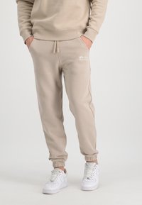 Beige bomulds sweatpants med elastiske manchetter og snor, der har et lille hvidt logo på venstre lommeområde. Bæres med hvide sneakers.