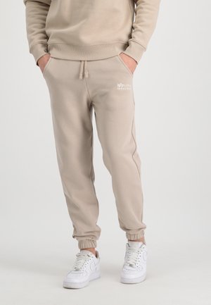 Beige Baumwoll-Sweatpants mit elastischen Bündchen und Kordelzug, mit einem kleinen weißen Logo im Bereich der linken Tasche. Getragen mit weißen Sneakers.