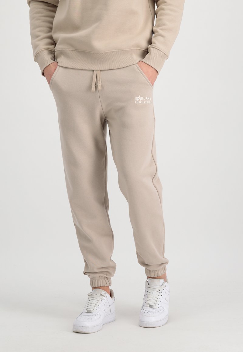 Beige bomulds sweatpants med elastiske manchetter og snor, der har et lille hvidt logo på venstre lommeområde. Bæres med hvide sneakers.