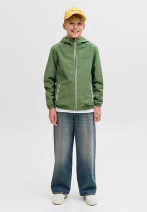 Usmívající se chlapec s žlutou čepicí, zelenou bundou na zip, volnými modrými džíny a bílými teniskami, stojící před jednoduchým světlým pozadím.