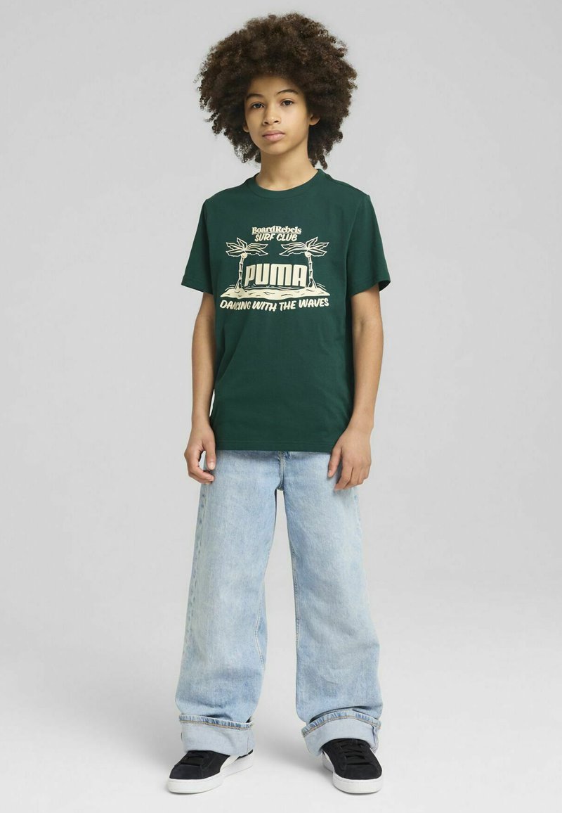 Camiseta de algodón verde con texto gráfico blanco y diseño de palmera. Combinada con unos vaqueros de pierna ancha de mezclilla azul claro y zapatillas negras.