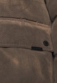 Brązowa kurtka puchowa o gładkiej, matowej fakturze. Posiada czarną naszywkę z logo oraz detal z guzikiem, ukazujące przeszycia i obfite wypełnienie.